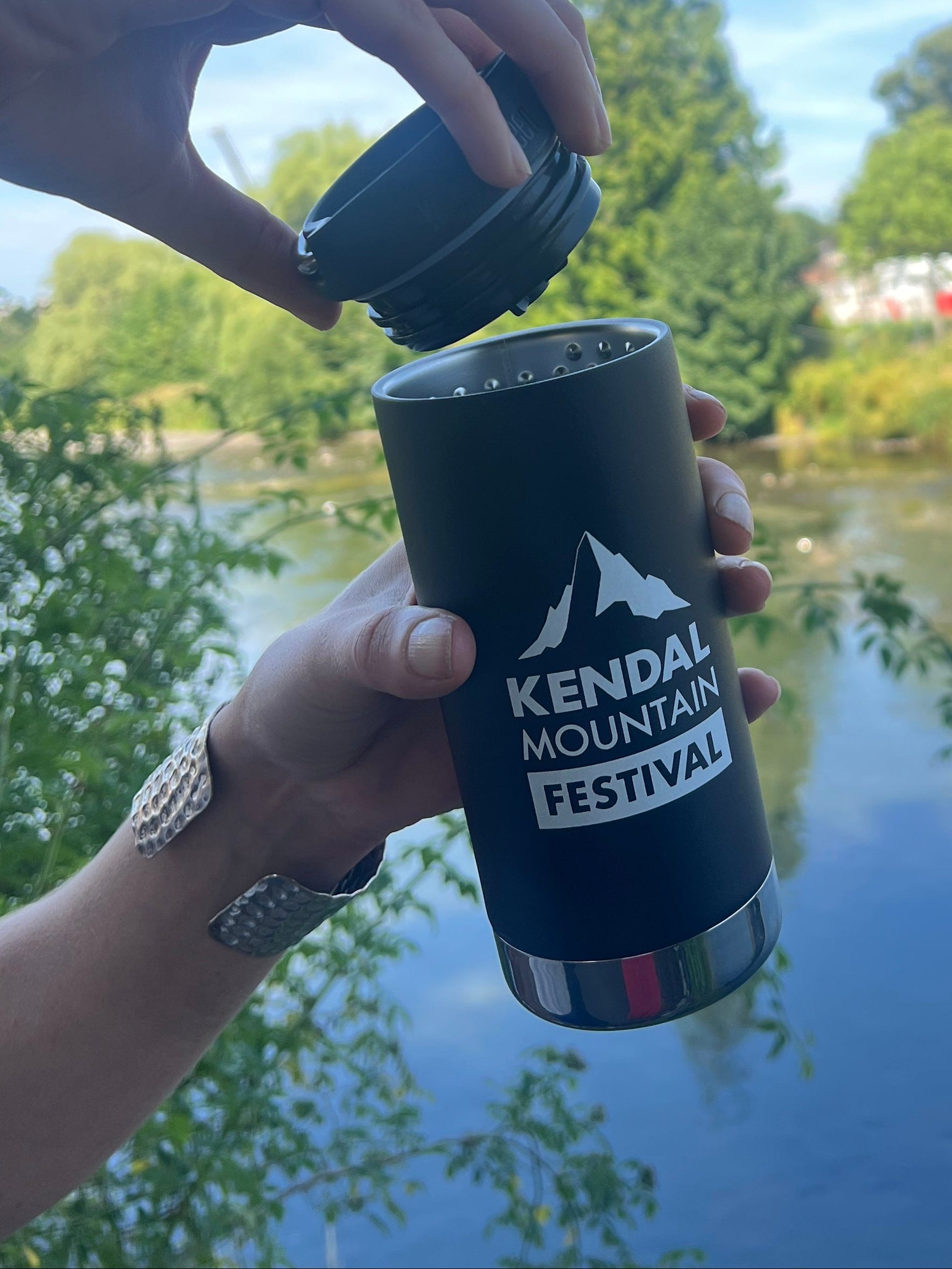A person outdoors by a lake holds a black insulated travel tumbler with the lid off, showing the white printed text 'KENDAL MOUNTAIN FESTIVAL' and mountain graphic. Trees, water, and blue sky are visible in the background.